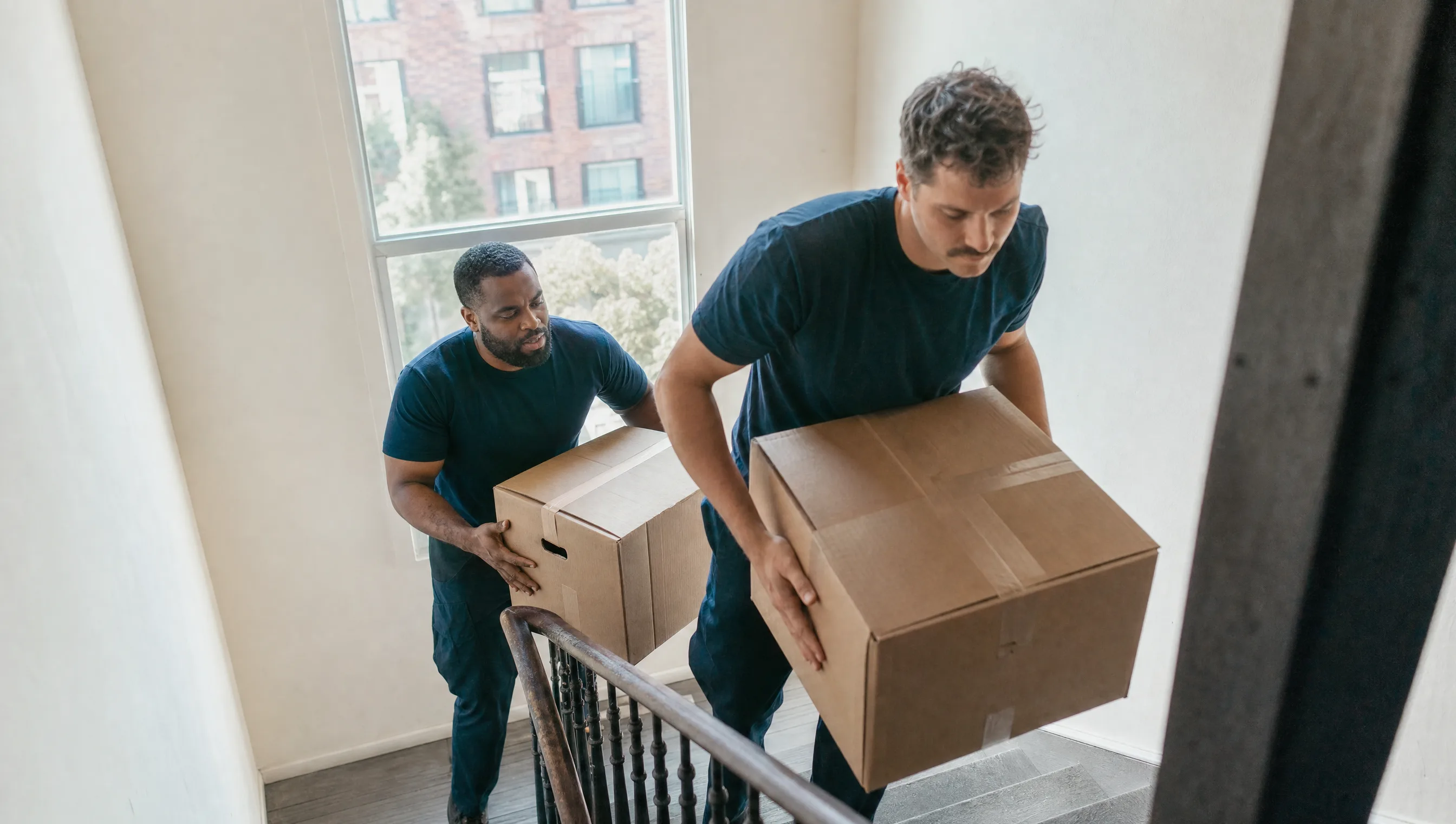 Two Triborough Trucking movers carrying cardboard boxes up a NYC walk-up staircase past a window